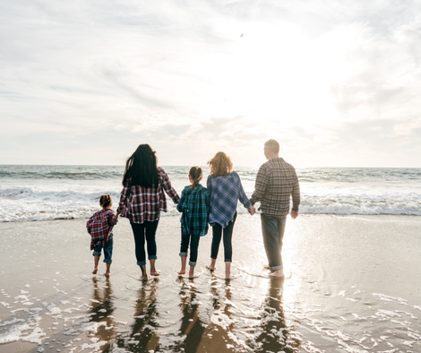 Family of five standing hand-in-hand in the shallow ocean waves at a beach, wearing casual plaid and colored clothing, viewed from behind as they look toward the horizon