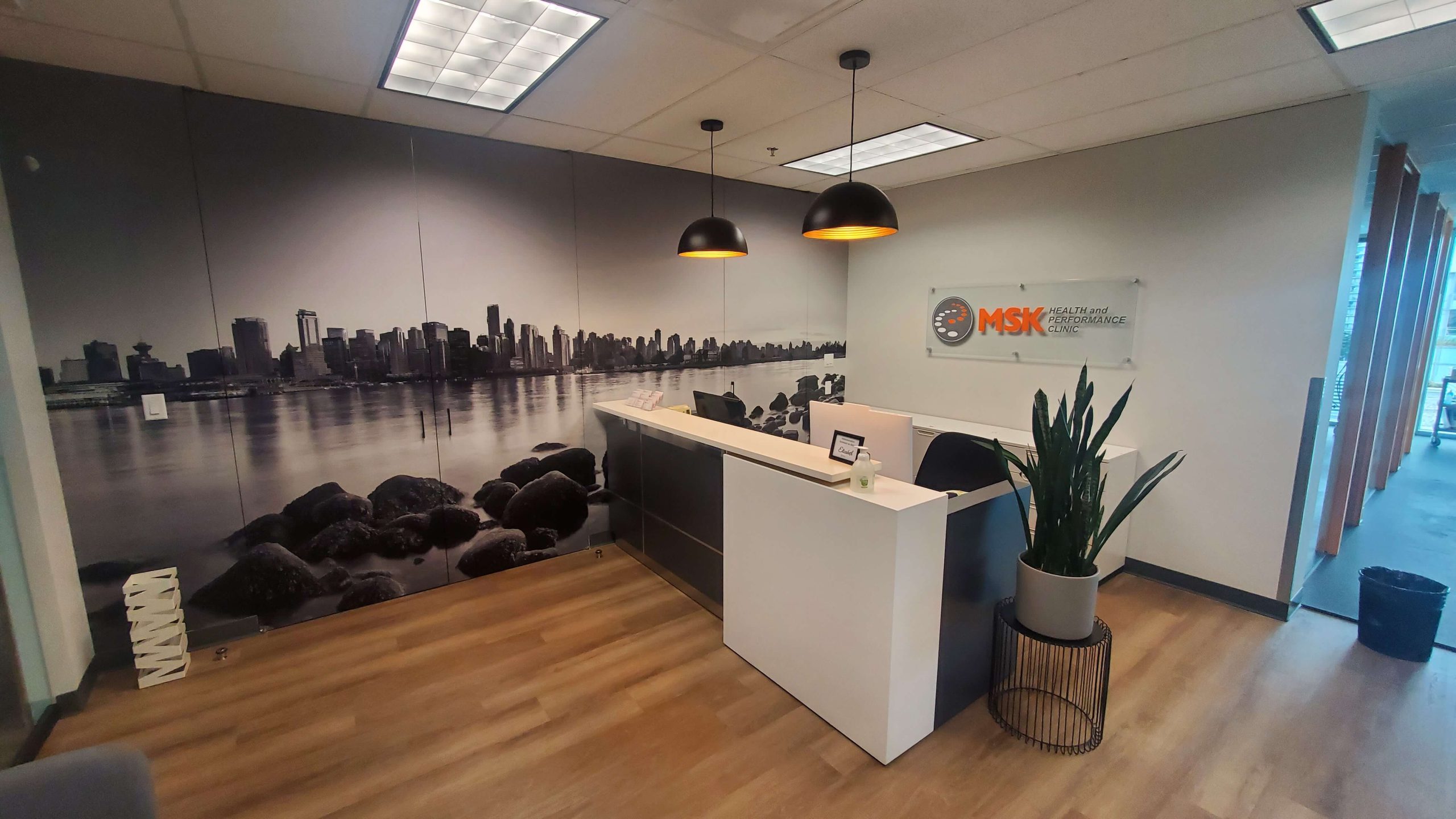 MSK Health and Performance Clinic reception area in Burnaby with modern white desk, pendant lighting, and cityscape wall mural