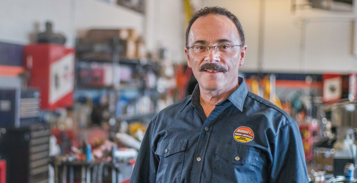 Portrait of a technician wearing glasses and a dark blue work shirt with Budget Brake & Muffler logo, standing in an automotive service shop in Burnaby