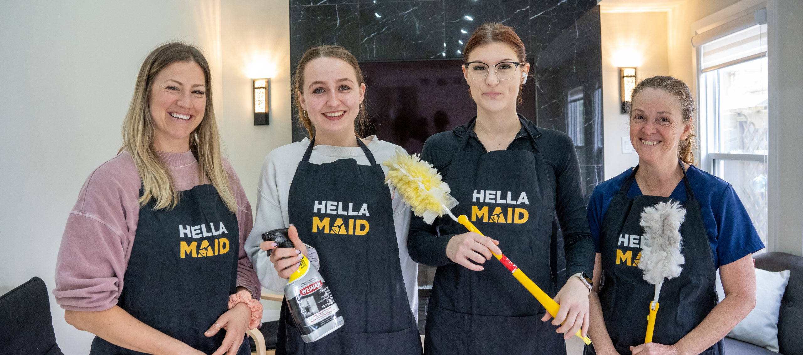 Four female cleaning service staff members wearing black Hella Maid aprons, holding cleaning supplies and smiling at the camera in a modern hallway