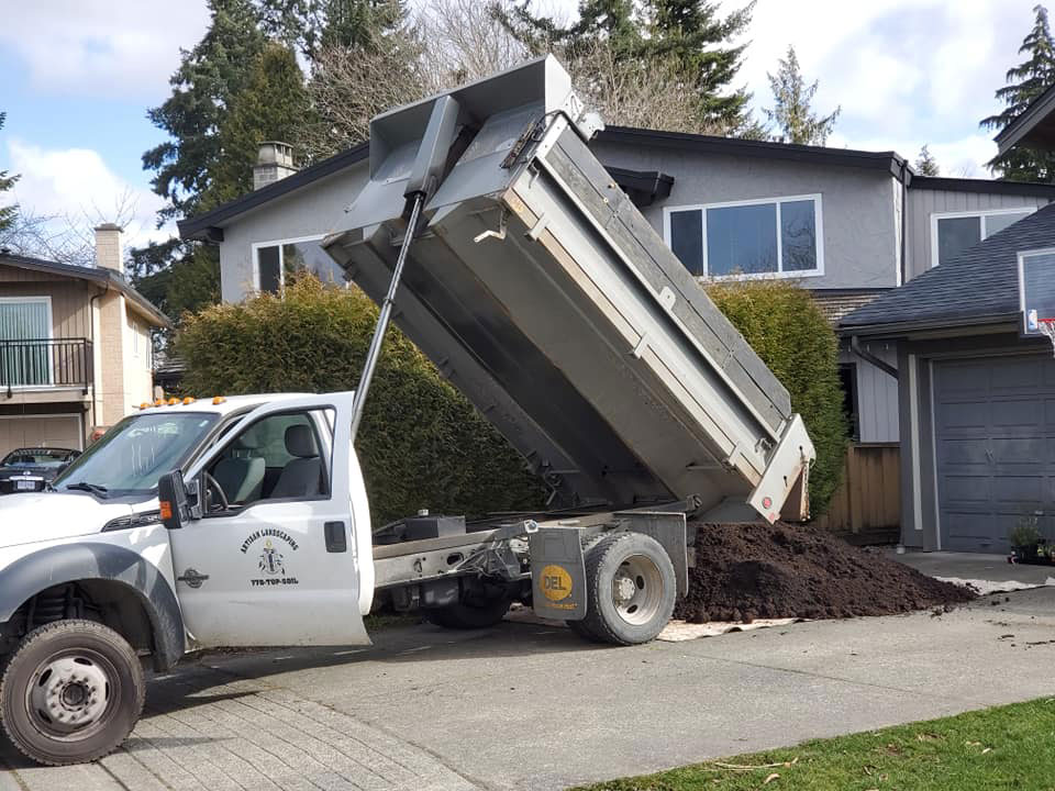 White dump truck with raised bed unloading dark soil or mulch in a residential driveway in Burnaby, with modern houses and evergreen trees in background