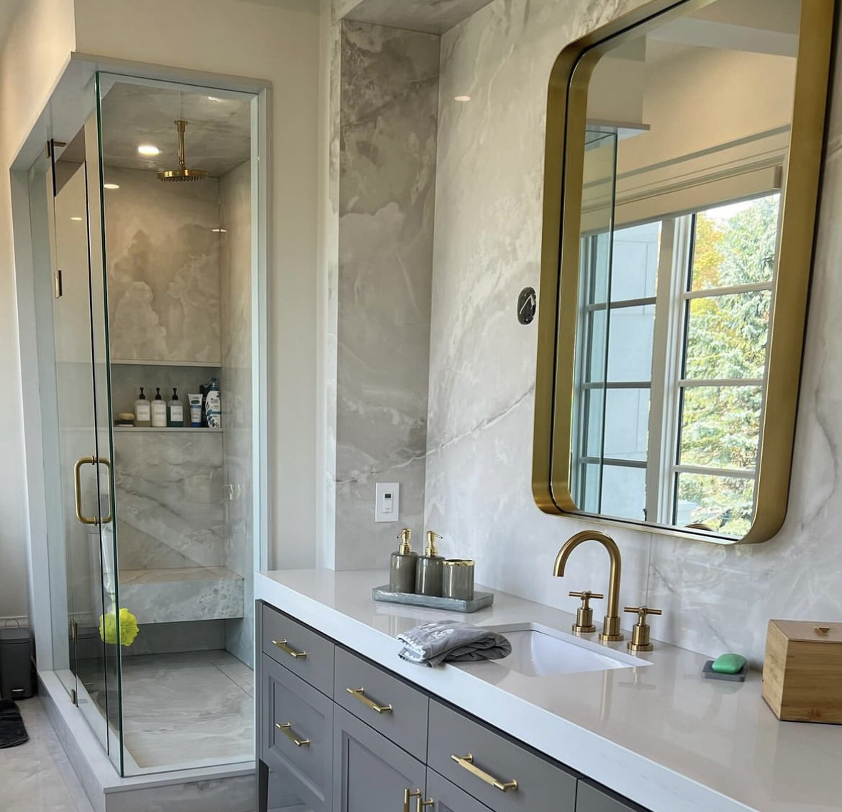 Modern bathroom in Burnaby featuring a glass-enclosed shower with marble tile, vanity with brass fixtures, and large gold-framed mirror