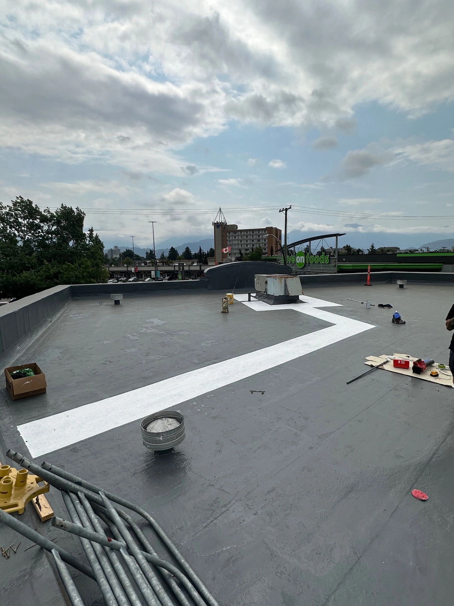 Flat commercial roof during construction or renovation in Burnaby with white membrane installation, tools and materials scattered across surface, mountains visible in background under cloudy sky