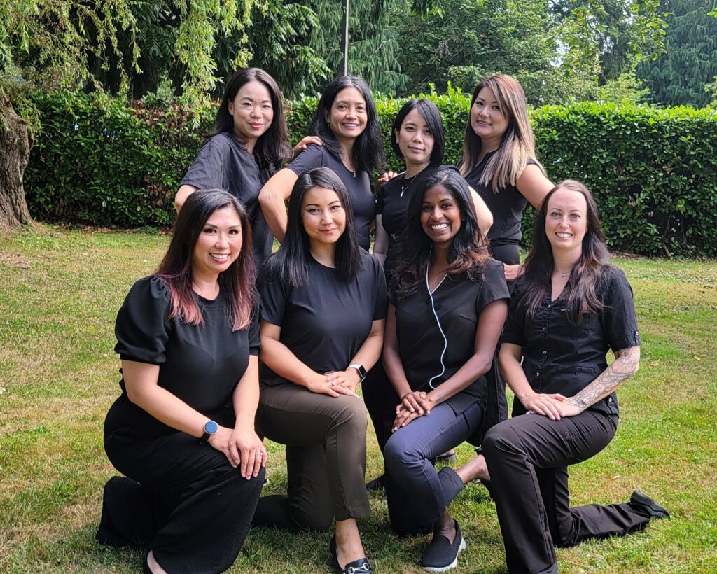My Smile Health Centre dental team in Burnaby - group photo of eight female dental professionals in black uniforms posing outdoors in a garden setting