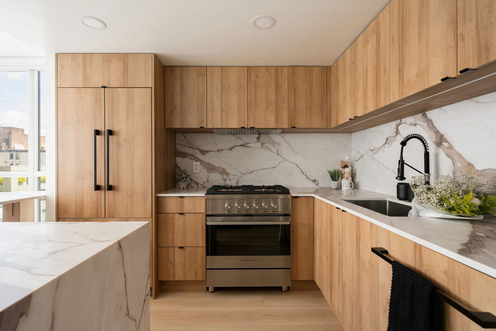 Modern kitchen with natural wood cabinetry, marble countertops and backsplash, stainless steel appliances, and black sink fixtures in a Burnaby home
