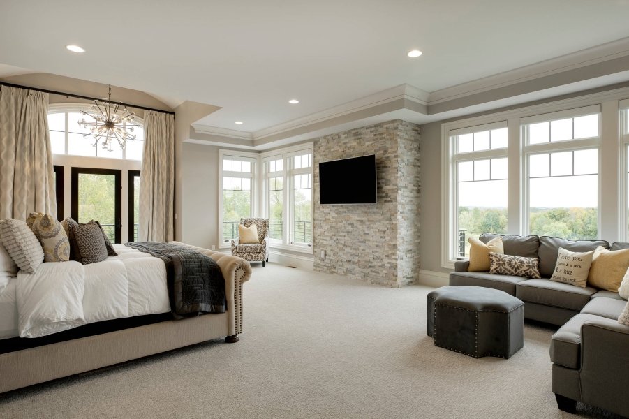 Luxurious master bedroom suite in Burnaby featuring a king bed with neutral bedding, sitting area with gray sofa, brick fireplace with mounted TV, multiple large windows with custom curtains, recessed lighting, and neutral color palette throughout