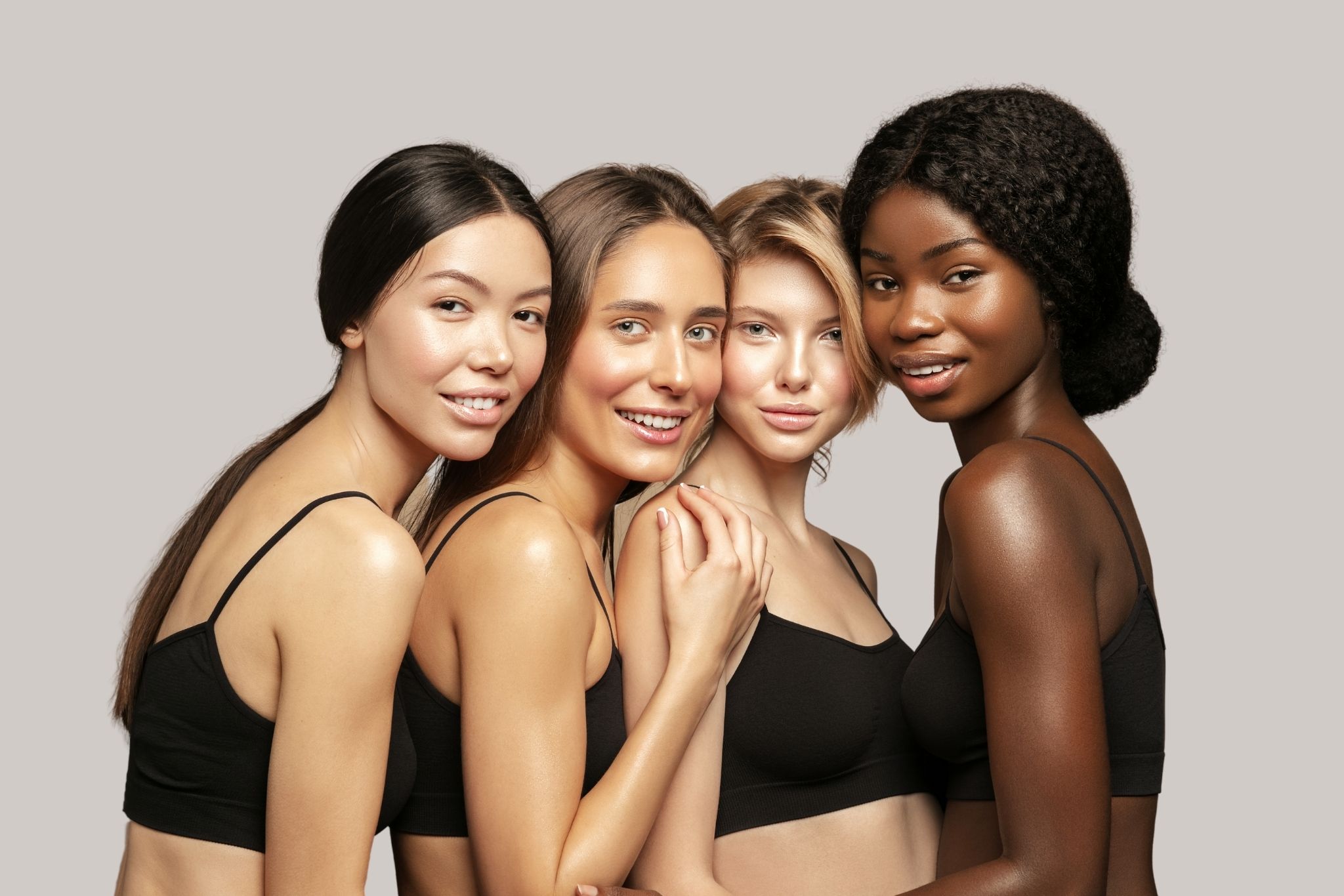 Four smiling women of different ethnicities wearing black sports bras posing together against a neutral gray background