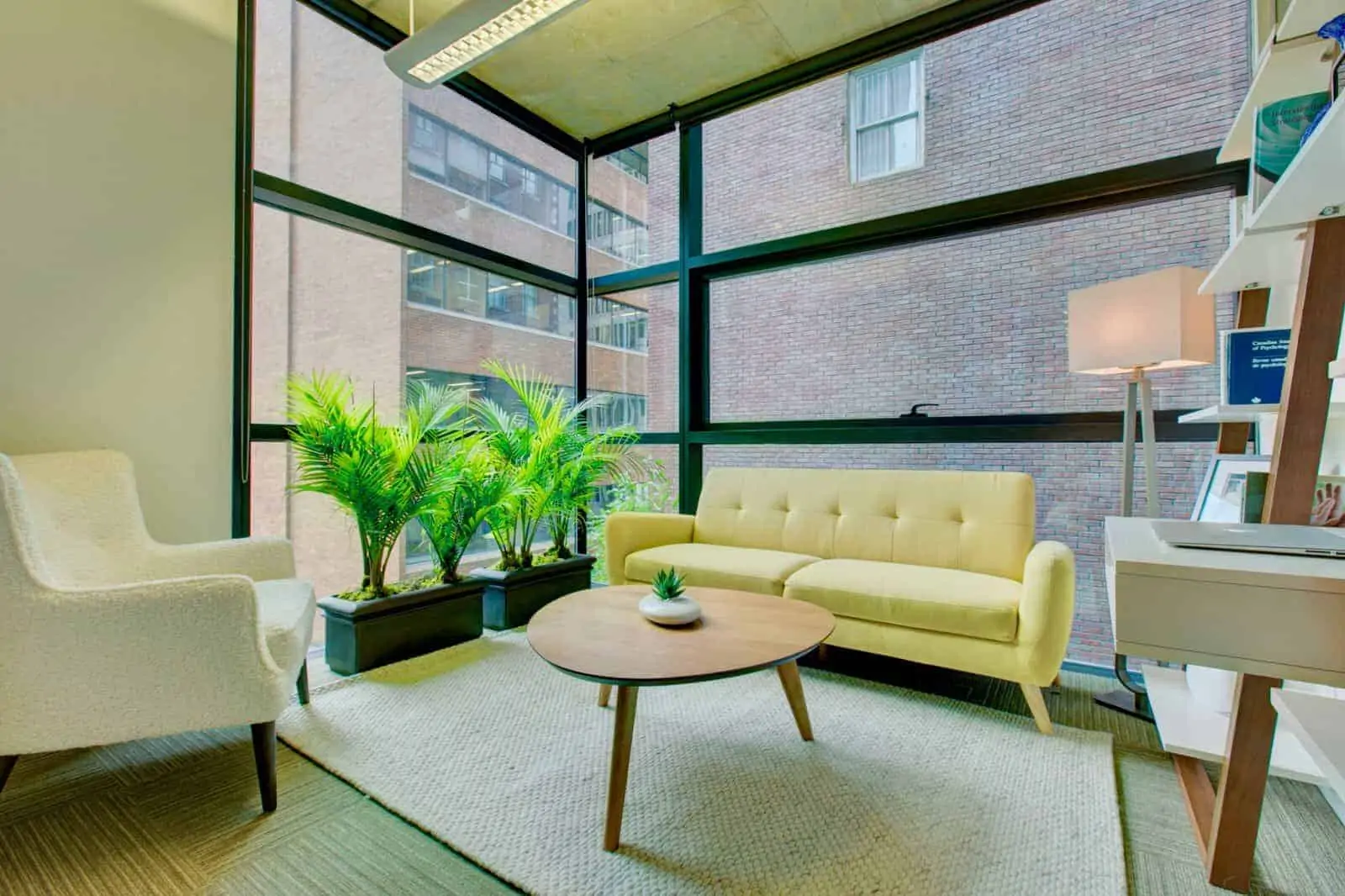 Modern counselling office in Burnaby featuring a bright seating area with a yellow sofa and cream armchair, potted green plants by large windows with green frames, a round wooden coffee table, and contemporary wooden desk