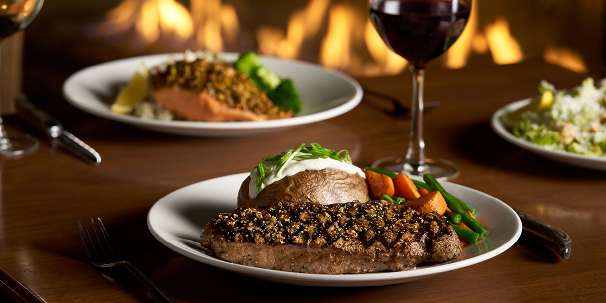 Gourmet steak dinner with herb crust, roasted vegetables, and sour cream topping served at The Keg Steakhouse + Bar in Burnaby, accompanied by red wine and side dishes
