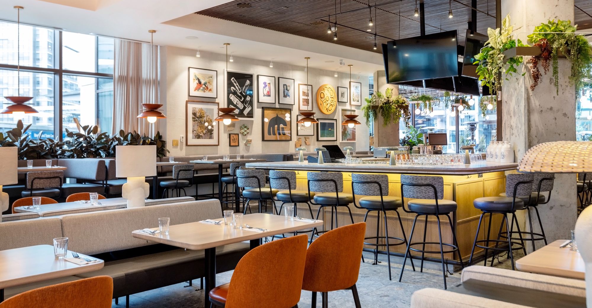 Earls Kitchen + Bar restaurant in Burnaby featuring a modern dining space with a yellow bar, orange chairs, framed artwork on walls, hanging plants, and floor-to-ceiling windows overlooking the city