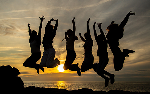 Six silhouetted people jumping joyfully against a golden sunset over the ocean, representing happiness and celebration in Burnaby