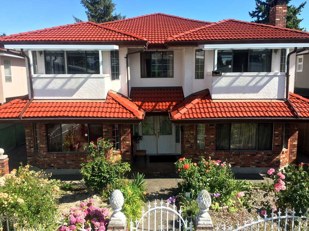 Modern two-story residential home in Burnaby with distinctive red tile roof, white upper level, red brick lower level, and manicured front garden with pink flowers and white gate