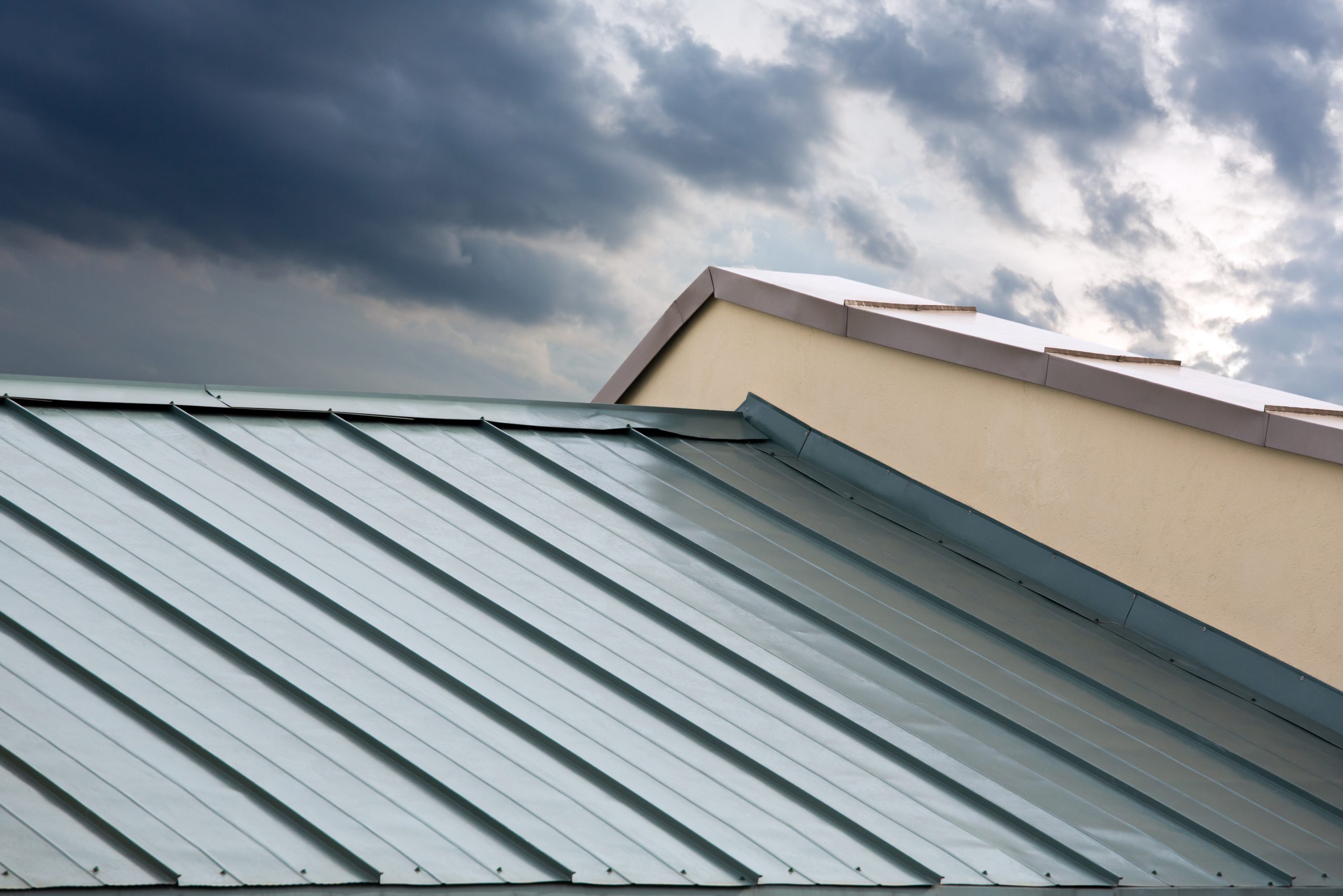 Close-up view of a metal standing seam roof with gray-blue panels alongside a cream-colored building wall against a cloudy sky in Burnaby