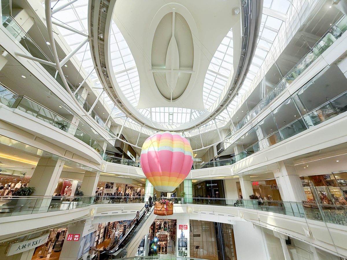 A colorful hot air balloon installation suspended in the center of Metrotown Mall in Burnaby, with multiple levels of retail stores and skylights visible above