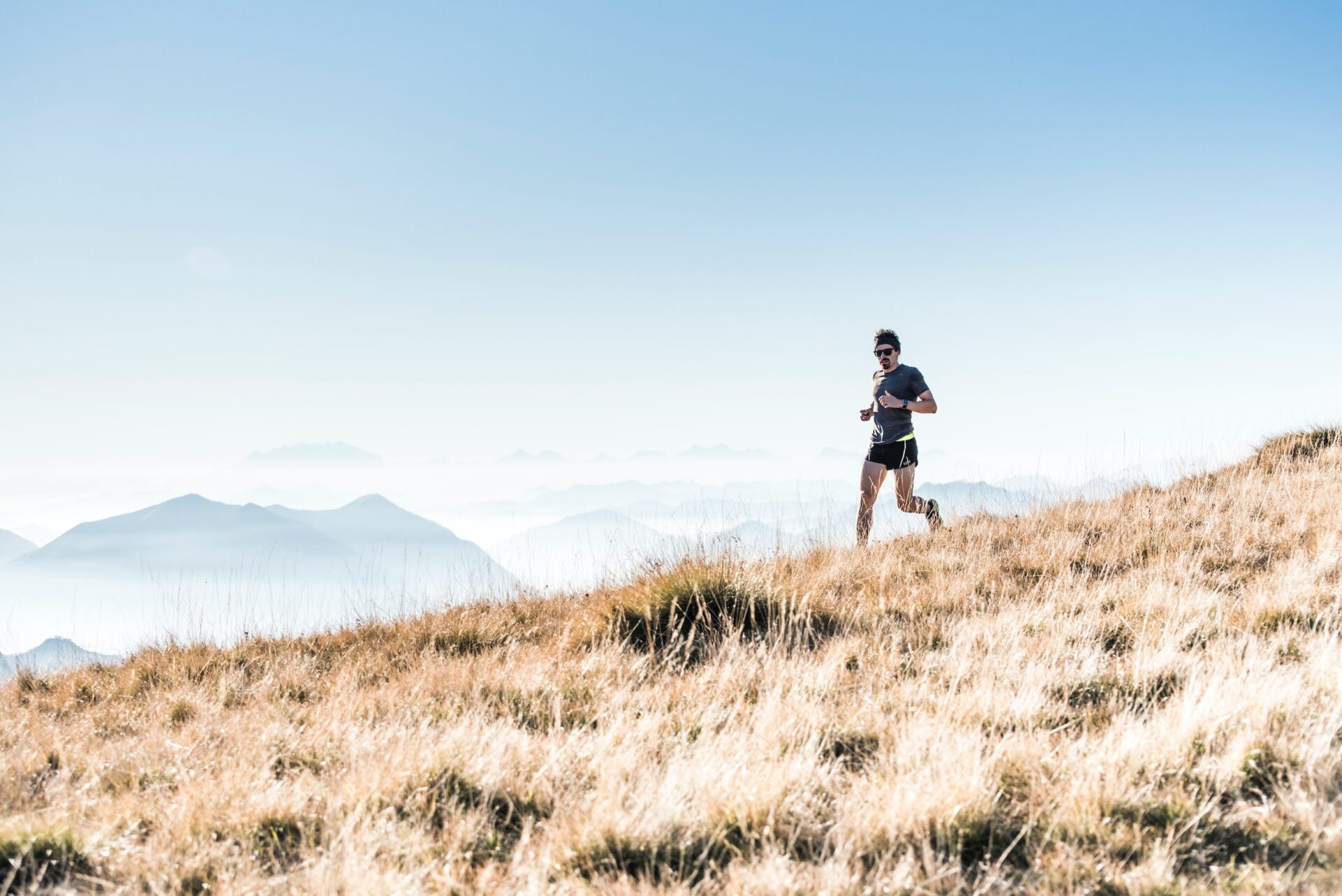 Male runner jogging uphill on a dry, grassy mountain slope with distant mountains visible in the background under clear blue sky
