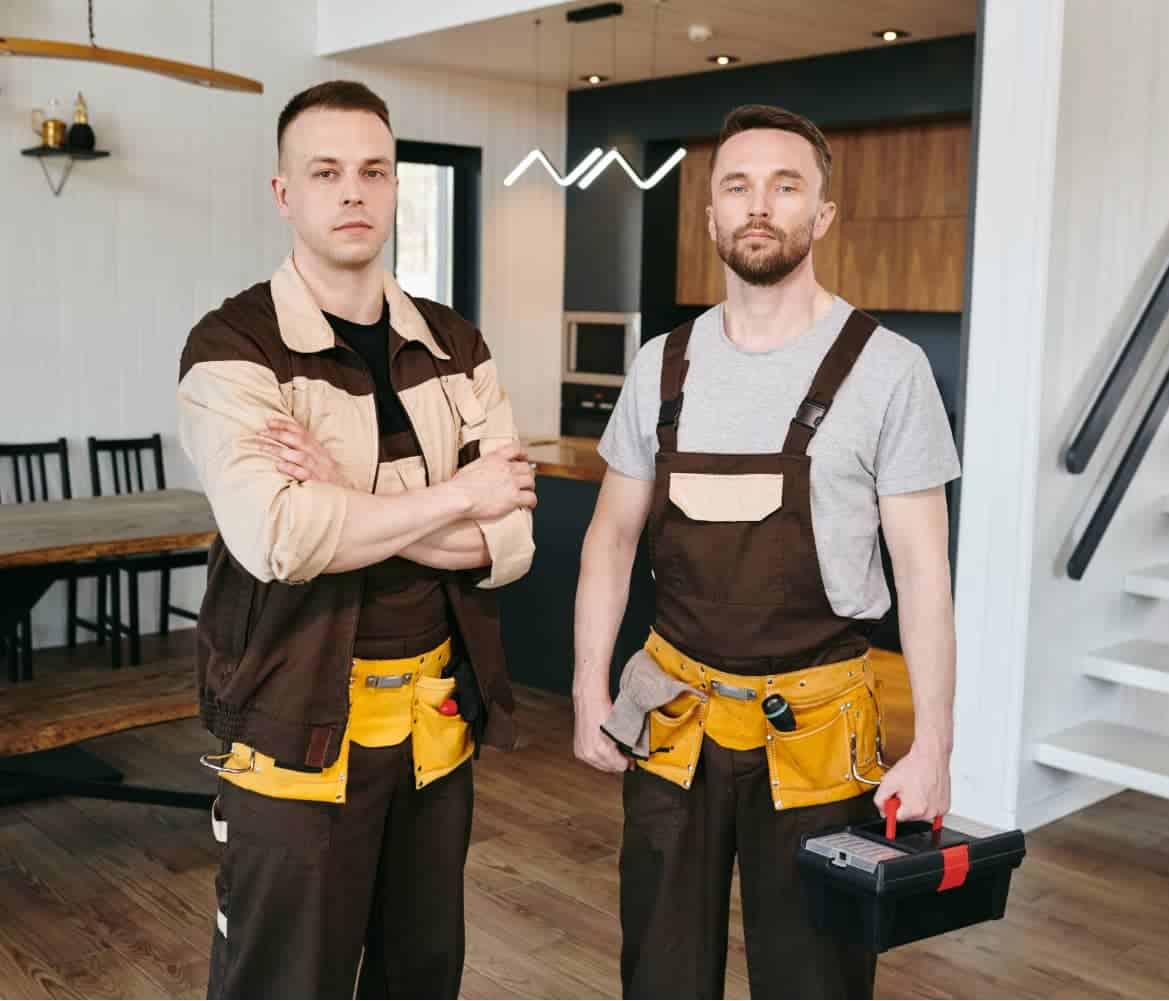 Two professional plumbers in brown and yellow work aprons standing in a modern kitchen renovation in Burnaby, BC, one holding a black and red toolbox