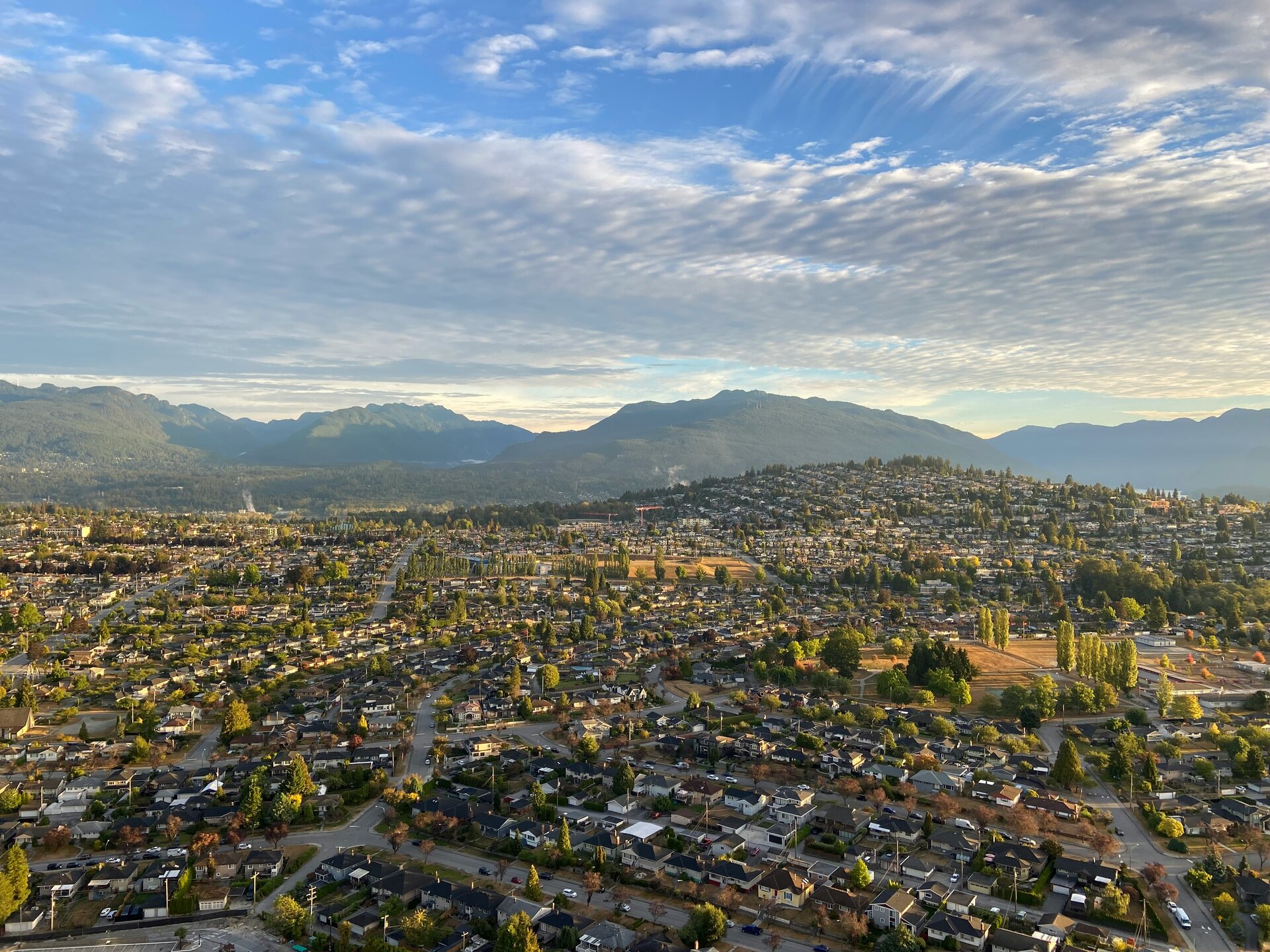 Aerial view of Burnaby, BC showing residential neighborhoods, tree-lined streets, and surrounding mountains under a blue sky with clouds