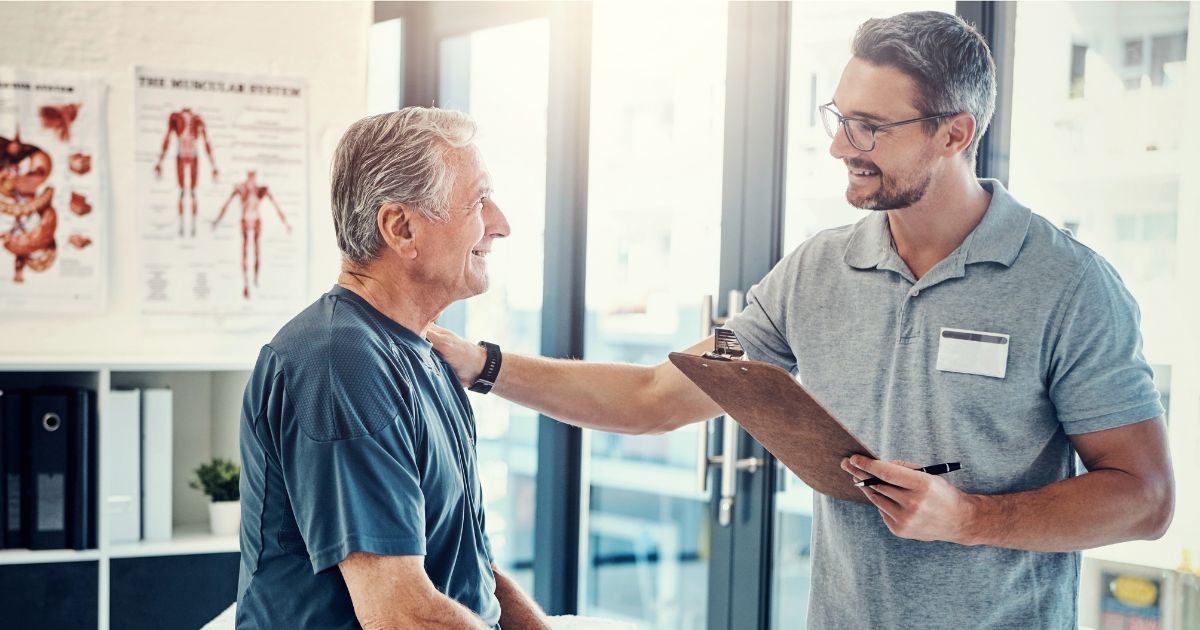 A smiling healthcare professional in a gray polo shirt with a name badge holding a clipboard, conducting a shoulder examination on an older male patient in a medical office setting with anatomical charts visible on the wall in Burnaby
