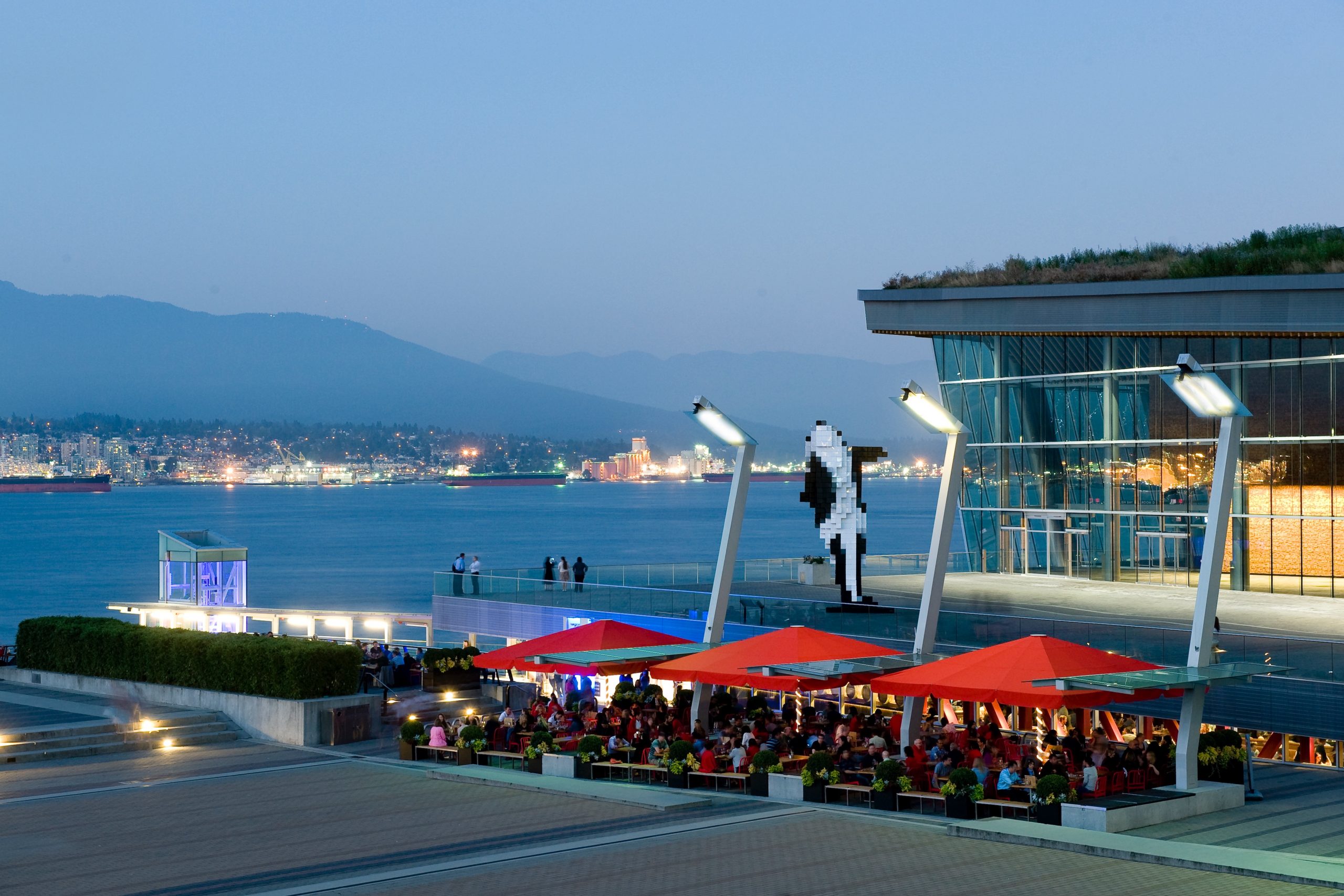 Waterfront dining area at Tap & Barrel in Brentwood, Burnaby, featuring red umbrellas, modern architecture, and views of the water and North Shore mountains at dusk