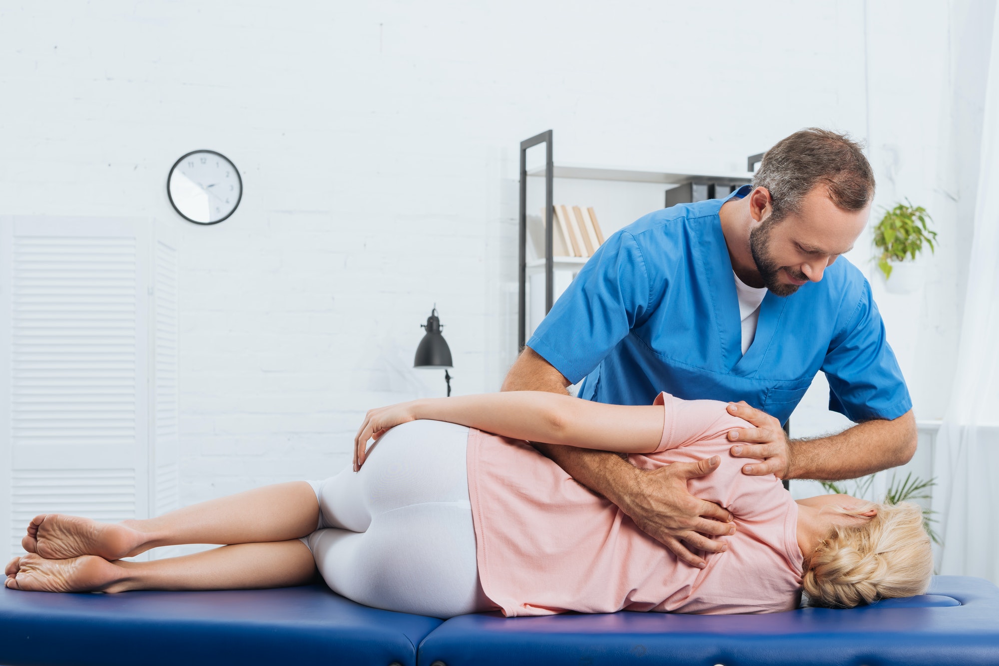 A physiotherapist in blue scrubs performing a spinal examination on a patient lying on a treatment table at a Burnaby physiotherapy clinic