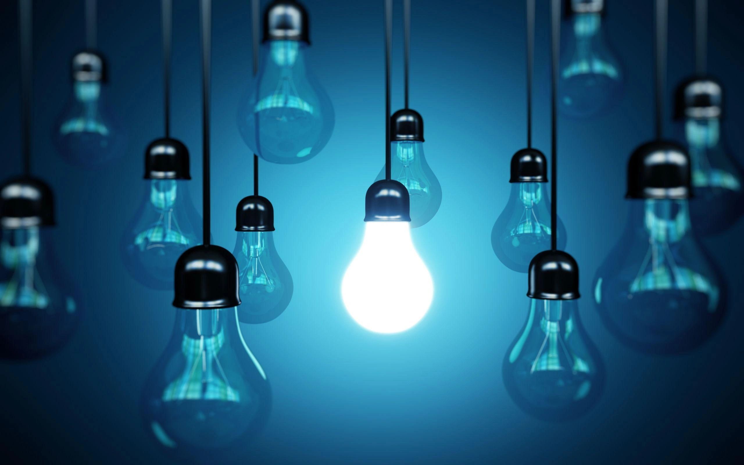 A bright white LED light bulb illuminated among dimmer traditional bulbs hanging against a blue background, representing innovation and energy efficiency for Main Street Electric Ltd. in Burnaby