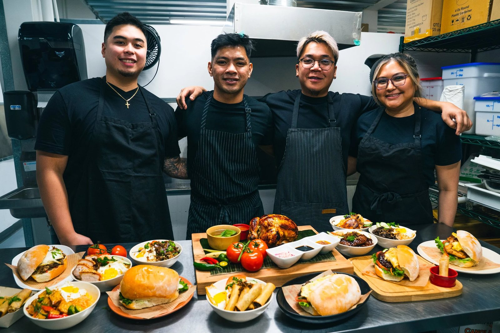 The four friends behind Halina in black aprons, standing in the kitchen behind a table of plated Filipino dishes